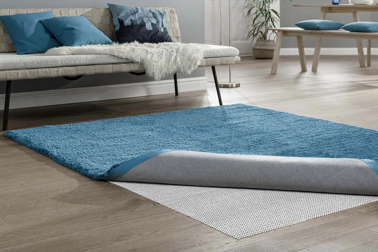 Illustration - Ihr Experte für schönes Wohnen - Ein Wohnzimmer in dem ein blauer Teppich liegt der umgeschlagen ist, die Teppichunterlage, die genau so groß ist, wie der Teppich, ist zu sehen.