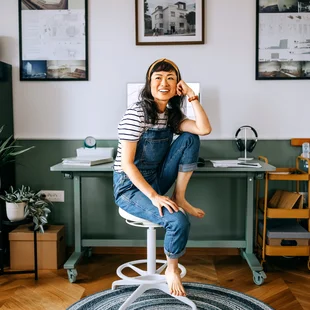 Illustration - Ihr Experte für schönes Wohnen - Porträt einer fröhlichen, lässig gekleideten schönen Frau, die am Schreibtisch im Homeoffice sitzt, unter ihrem Stuhl liegt ein runder Teppich.