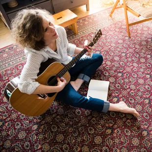 Illustration - Ihr Experte für schönes Wohnen - Junge Frau sitze im Wohnzimmer auf dem Teppich und spielt Gitarre.